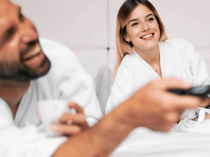 A couple in white bathrobes watching TV in a hotel bed.