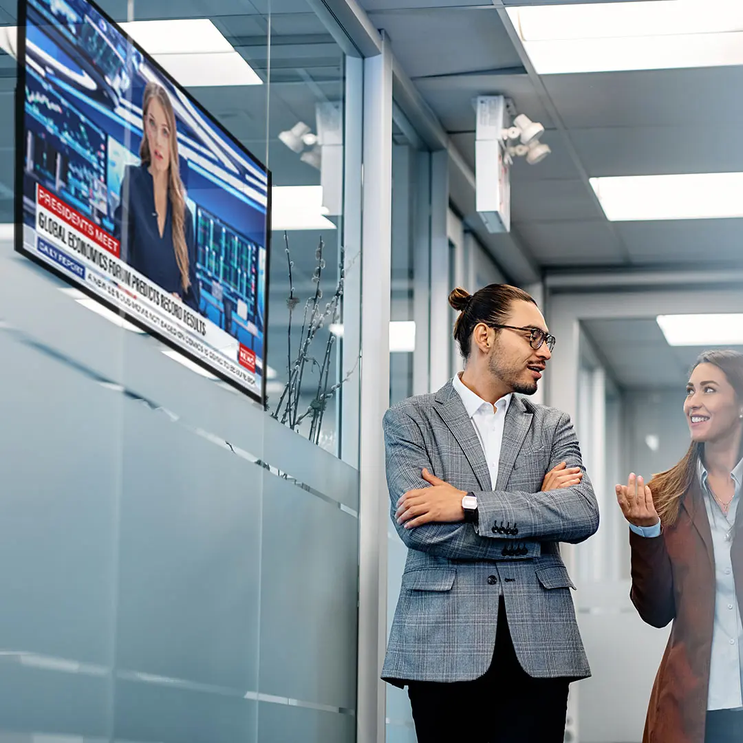Two colleagues walk past a TV screen showing office news. Två kollegor går förbi en TV-skärm som visar nyheter på kontoret.