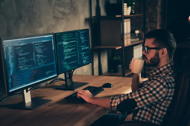 A coder with a coffee cup at computer screens. En kodare med kaffekopp vid datorskärmar.