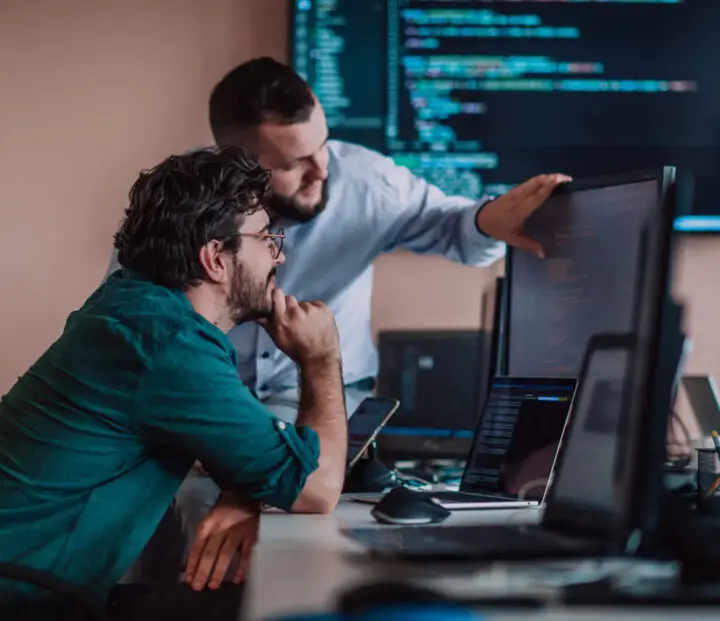 Two men are looking at a computer screen displaying code, with several computers nearby.