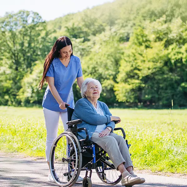 A carer pushes an elderly woman in a wheelchair through a green and sunny landscape