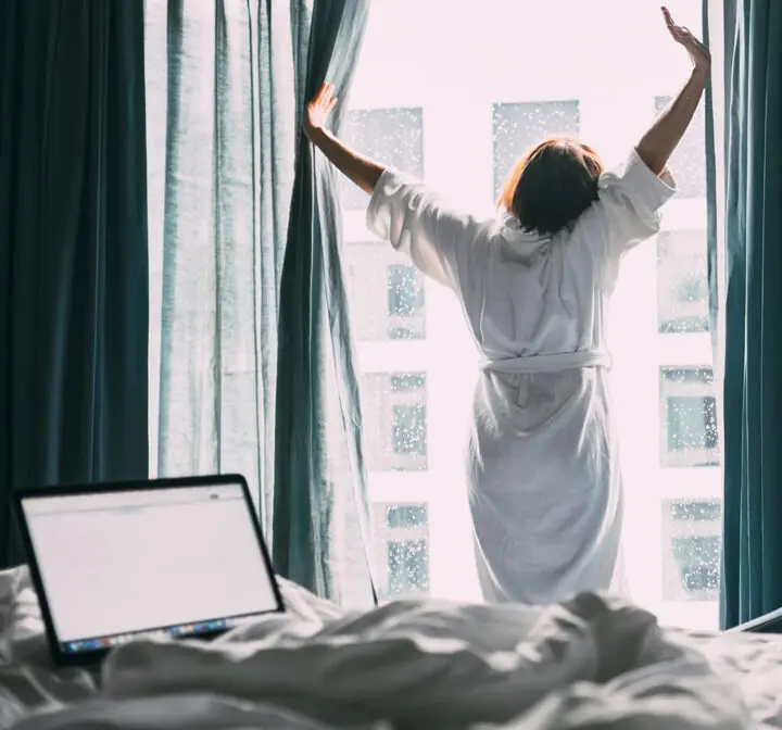 A woman in a dressing gown stretches and looks out of a hotel window, her laptop open on the bed