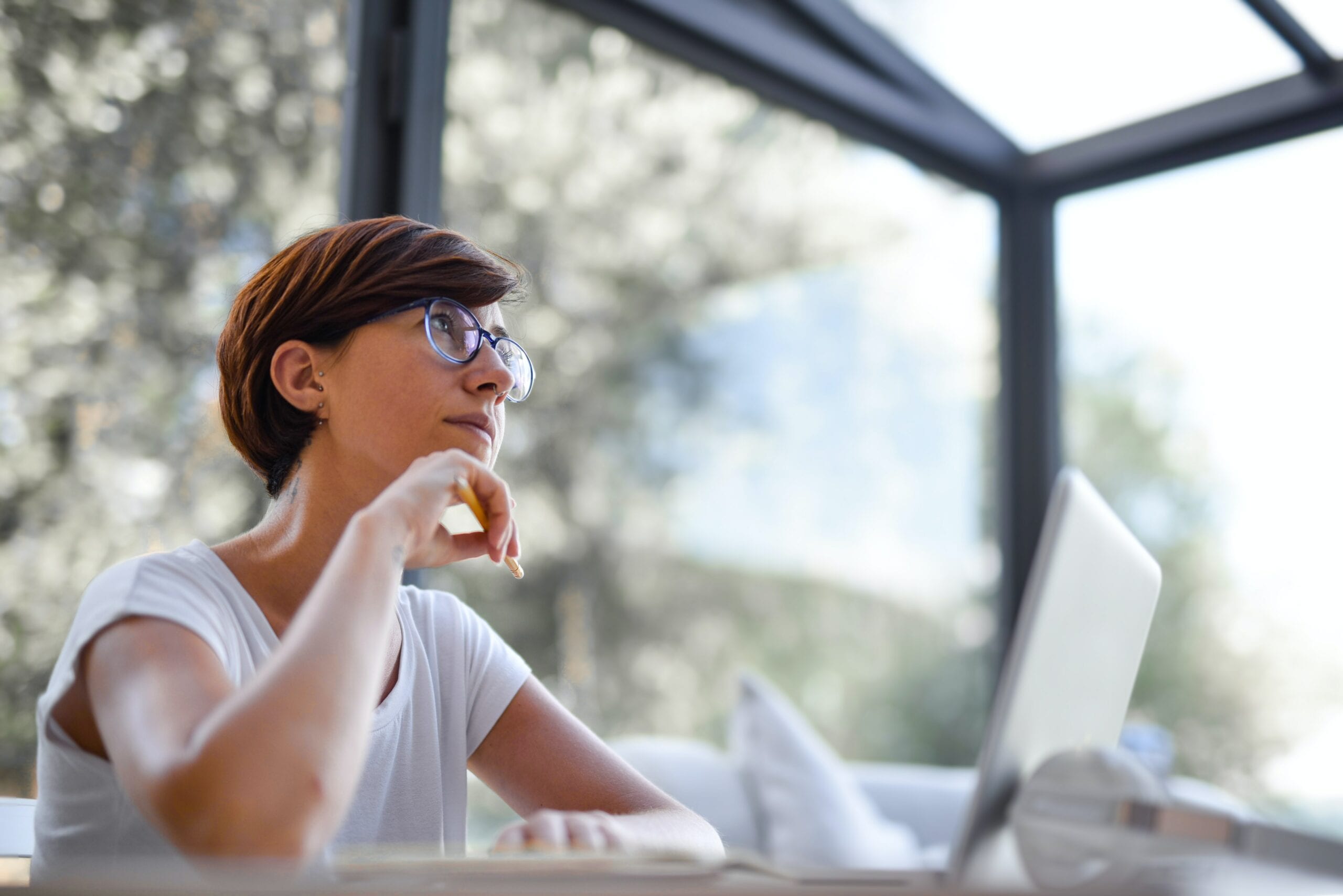 Woman working at laptop in front of large glass walls with foliage outside.