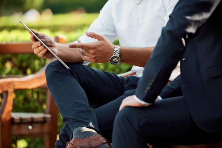 Businessmen sitting on a park bench discussing something on a screen. Affärsmän sitter på en parkbänk och diskuterar något på en skärm.