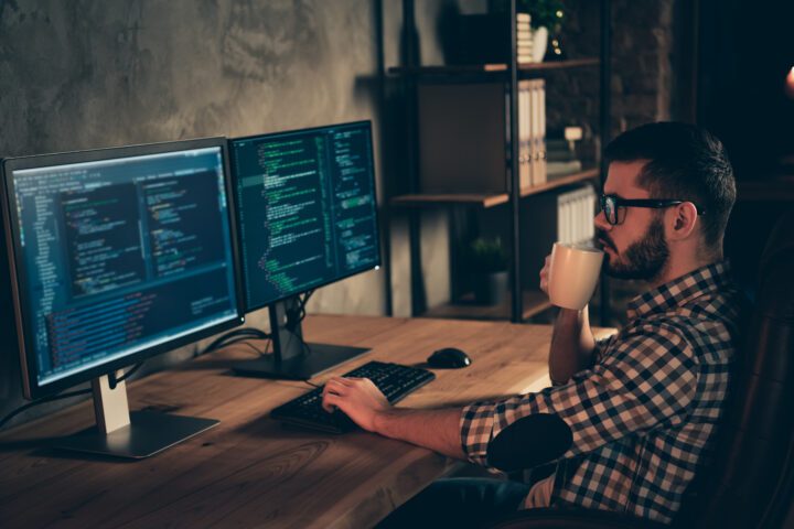 A coder with a coffee cup at computer screens in a dark room.