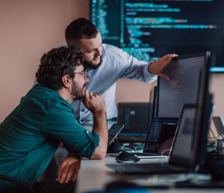Two men are looking at a computer screen displaying code, with several computers nearby.