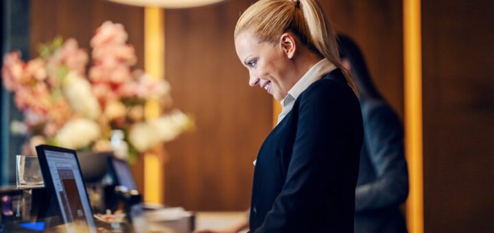 A cheerful hotel receptionist stands at the reception desk and works at the computer.