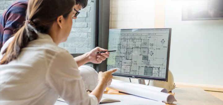Two colleagues are working at a screen with a building plan. Two hard hats and rolled-up papers are on the table.