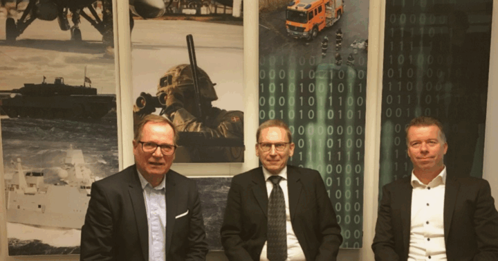 Three men in suits in front of a wall with military pictures