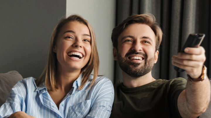 A happy couple watches TV in a hotel room and changes channels with the remote control.