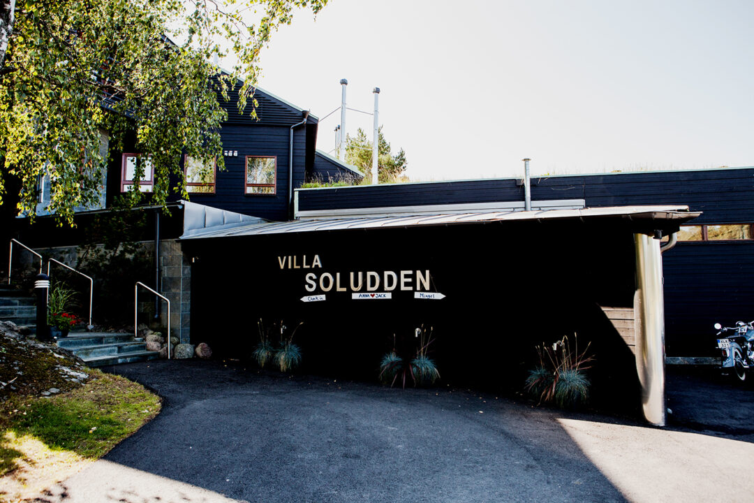 Svart trebygning med skiltet "Villa Soludden" ved inngangsparti og noen planter foran bygget.