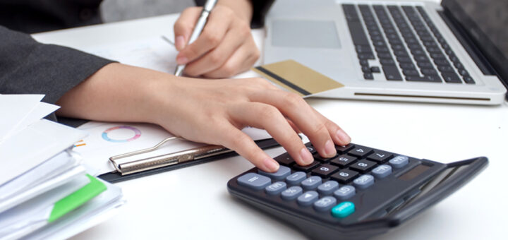 Person using a calculator and taking notes on a piece of paper, with a credit card, a laptop and a stack of papers also visible in the image.