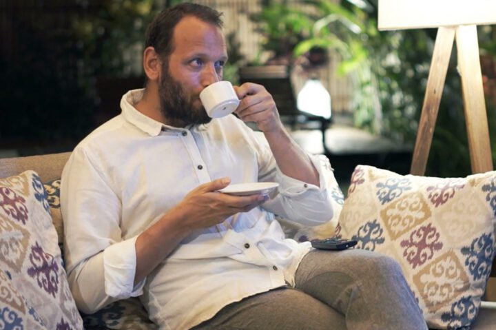 A person is sitting on a sofa, drinking coffee, and watching television.