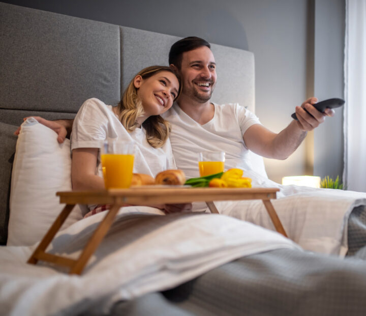 A couple watching TV in a hotel bed with breakfast in front of them