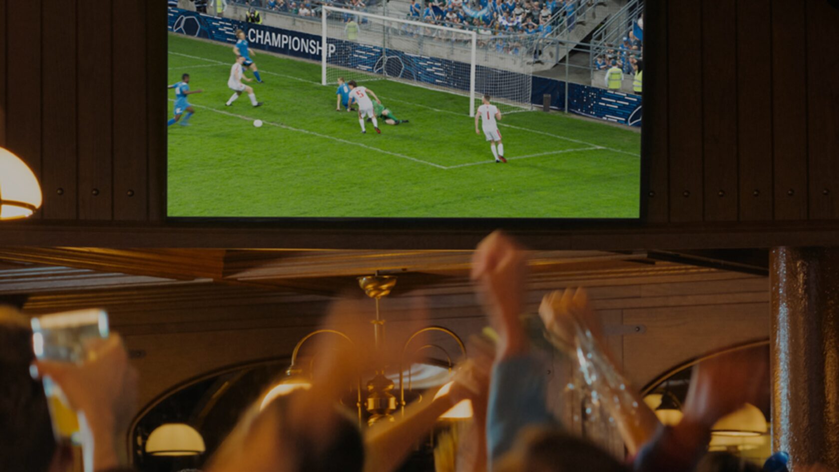 Football fans watch a match on a large screen in a warmly lit pub.