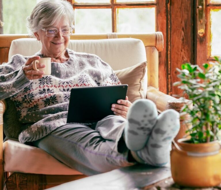 Eldre kvinne med grått hår sitter i stol, drikker kaffe og bruker nettbrett, ser avslappet ut.