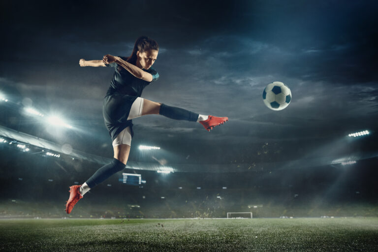 Female footballer kicking a ball in a stadium at night, with bright stadium lights in the background.