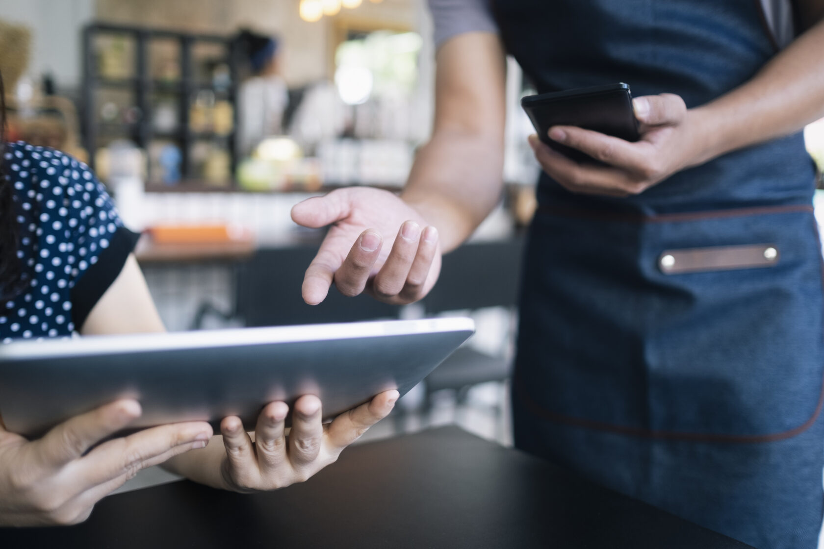 Two people in a restaurant, one showing something on a tablet, the other holding a mobile phone.