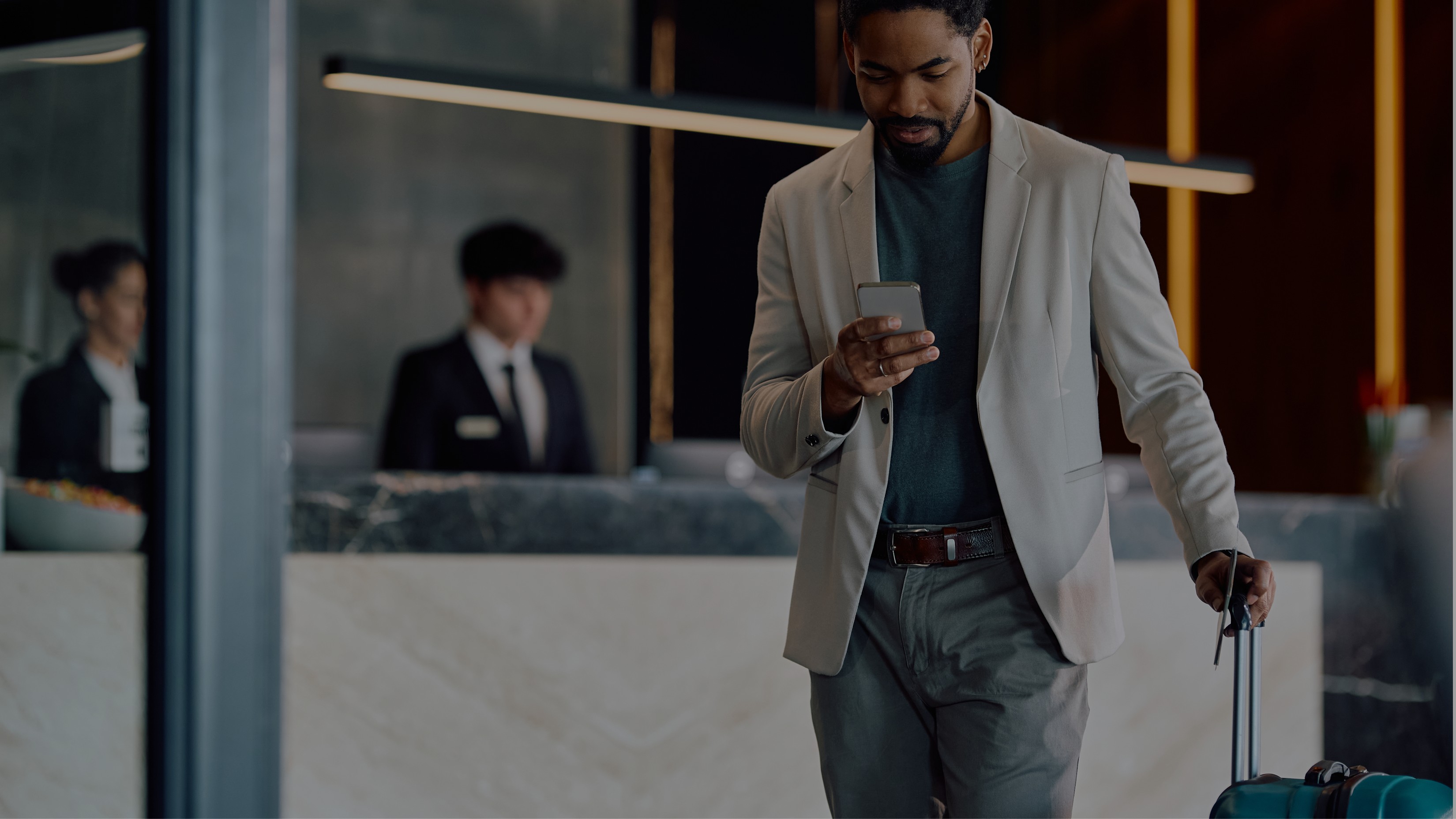Mann med blazer sjekker mobilen mens han står ved en koffert i hotellobbyen.