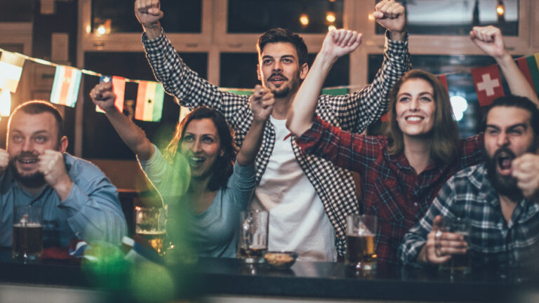 Five people at a bar are cheering in front of a TV, with beer glasses on the table and flags in the background.