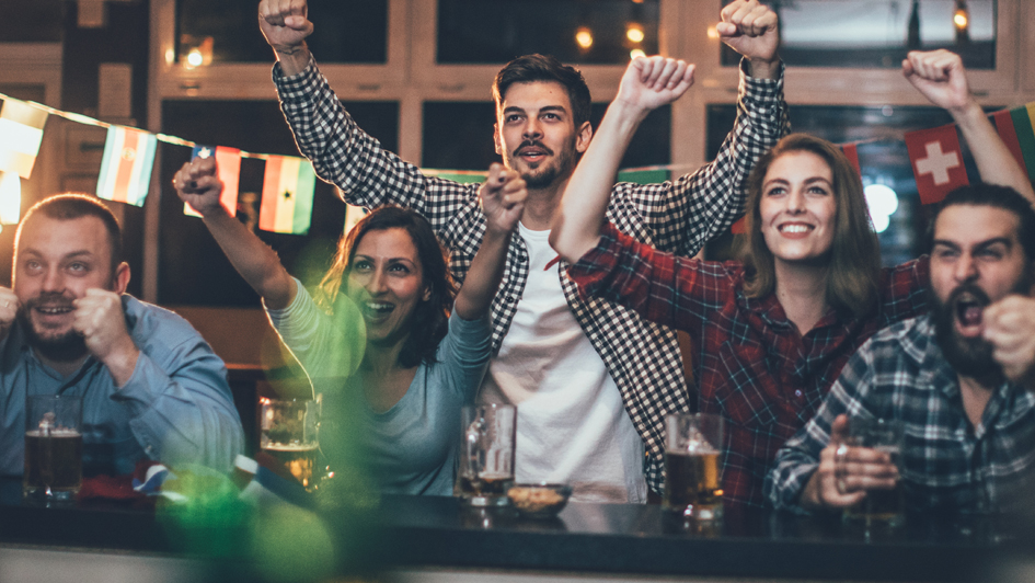 Five people at a bar cheer in front of the TV, with beer glasses on the table and flags in the background.