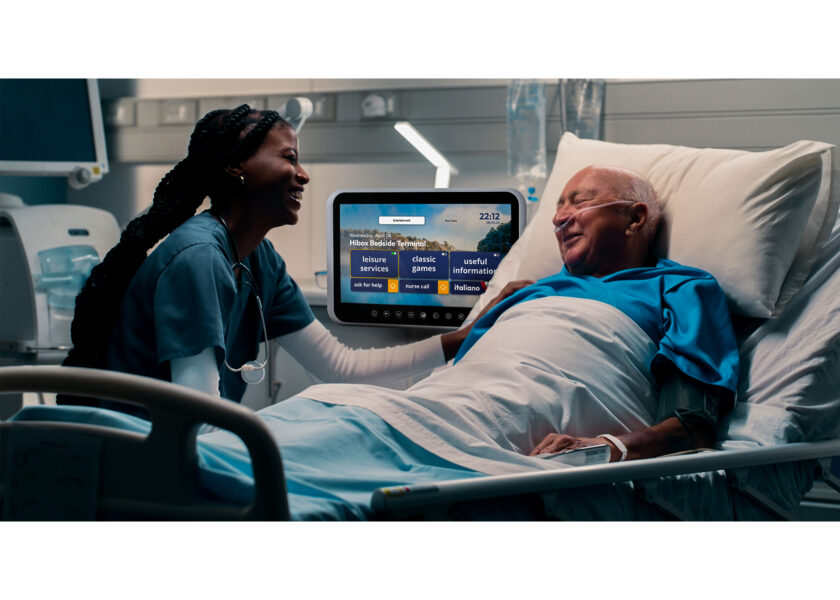 Nurse and elderly patient smiling in hospital room with a bedside terminal screen displaying service options.