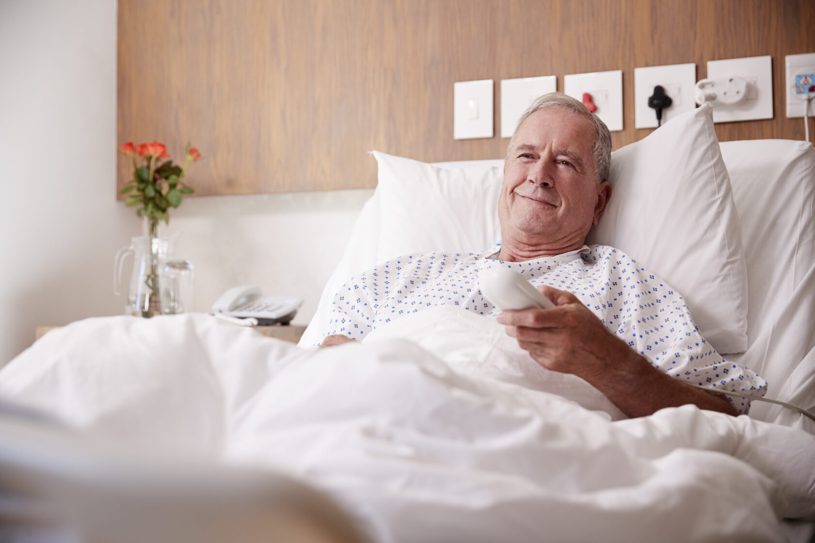 Older man in hospital bed holding a remote control and smiling, flowers and watering can in the background.
