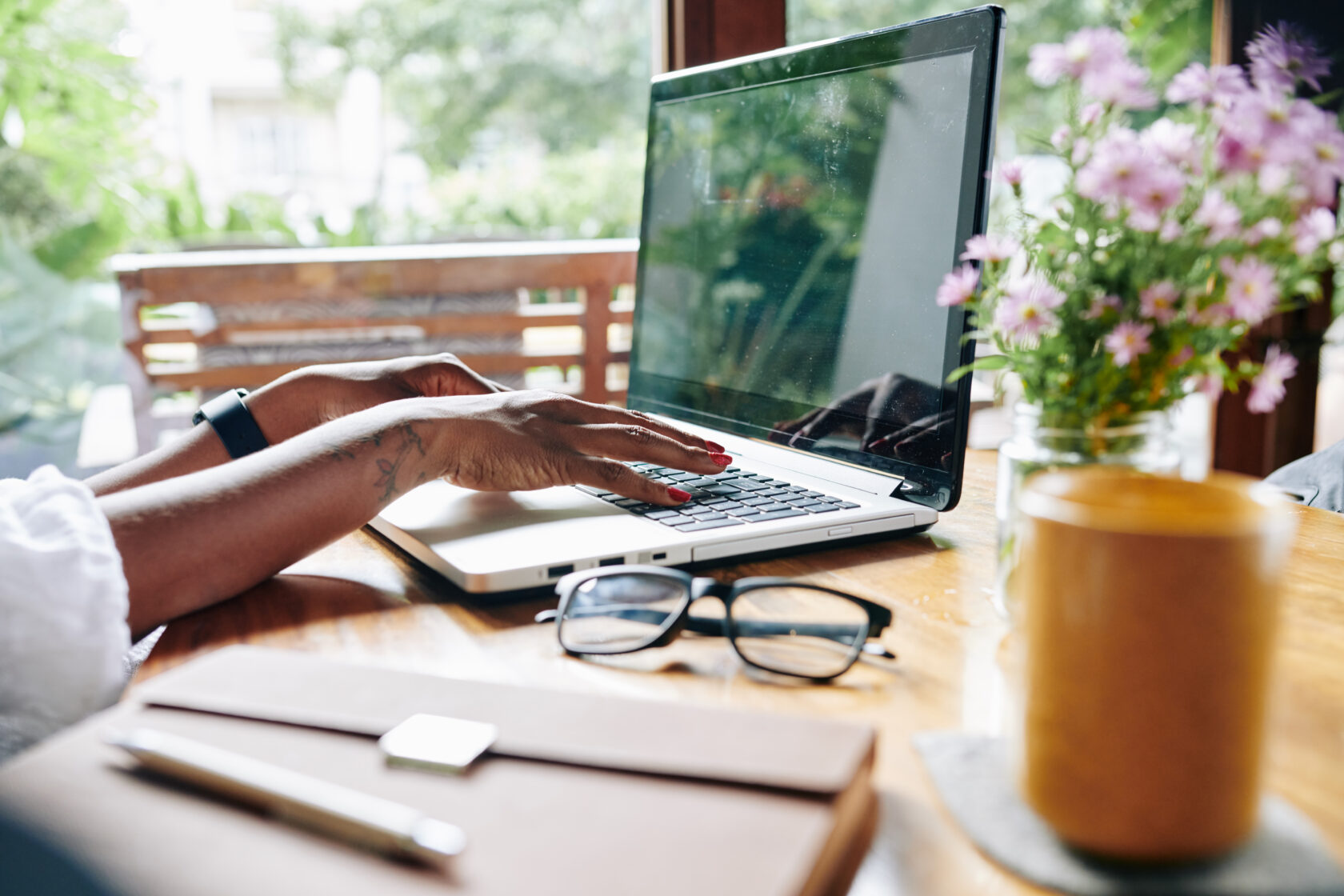 Person writing on laptop at a wooden table with glasses, coffee, notebook, and flowers beside.