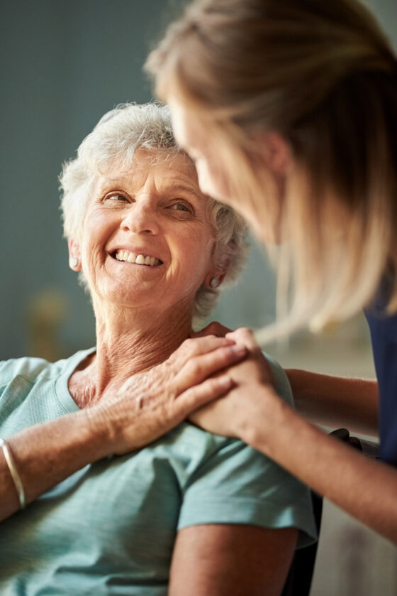 Smiling elderly woman receiving support from a younger woman holding her shoulders, both looking happy.