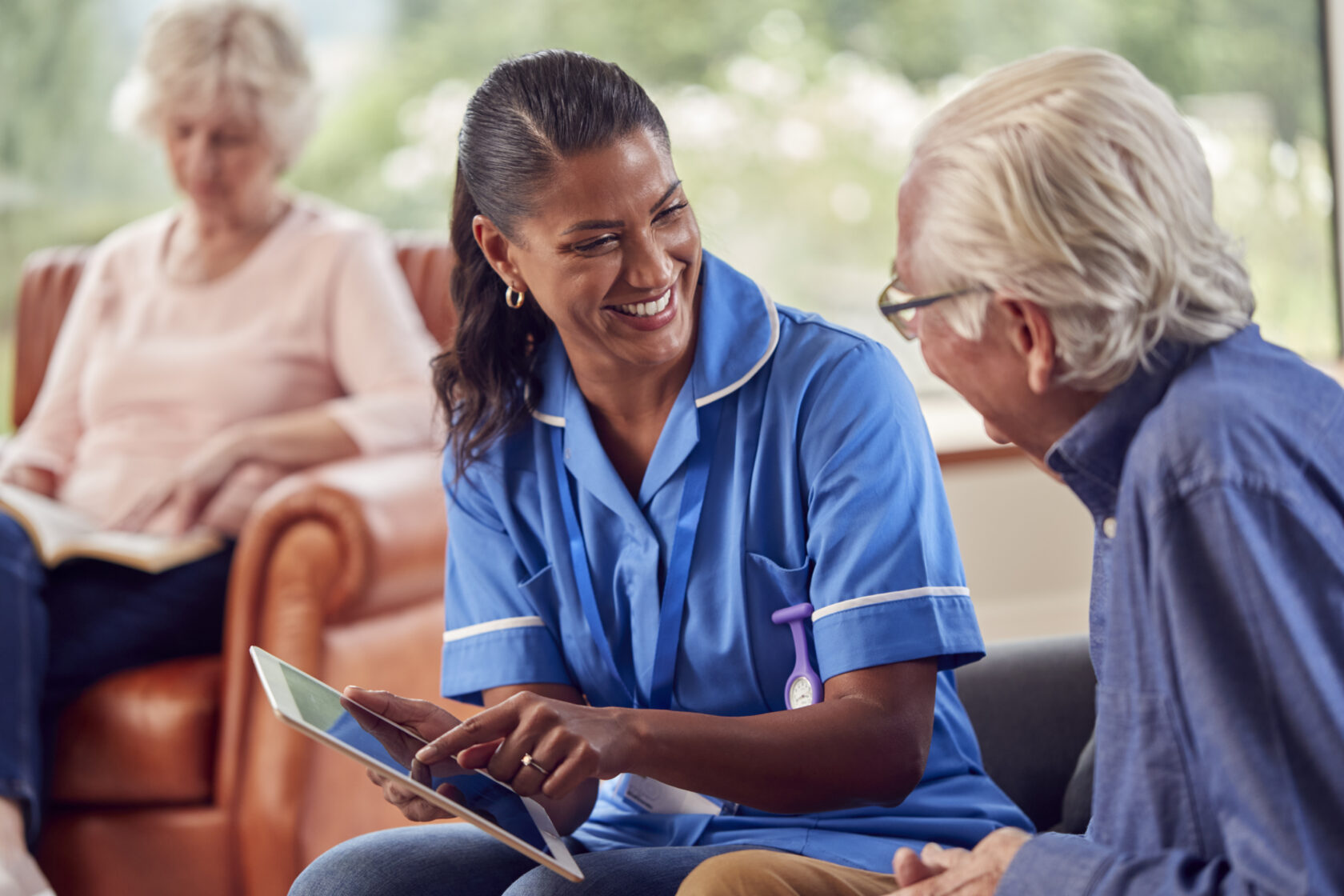 A nurse in blue uniform shows a tablet to an elderly man, both smiling. An elderly woman is sitting in the background.