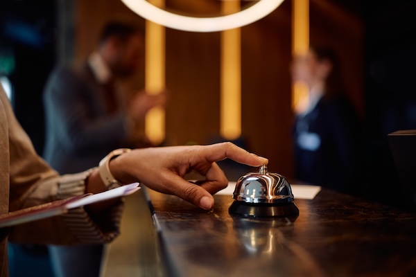 En hand trycker på en hotellreceptionens ringklocka, suddiga personer i bakgrunden.