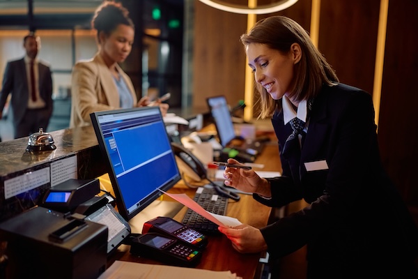 Kvinnlig receptionist arbetar vid dator på hotell, skriver ut papper, gäst checkar in i bakgrunden.