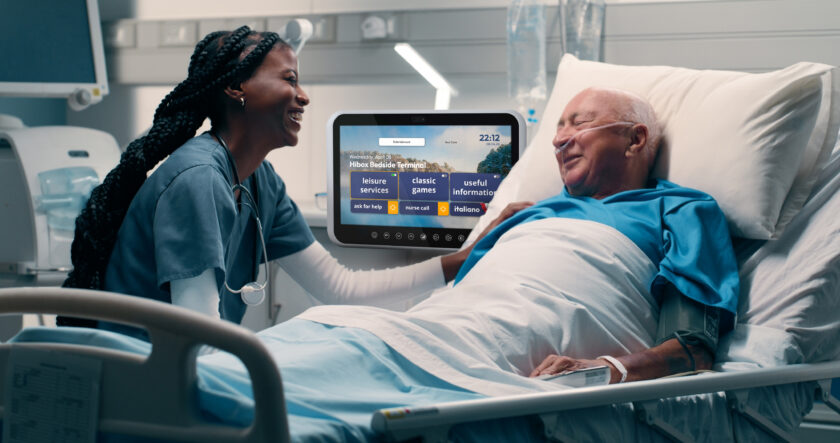 Nurse smiling and talking to an elderly man lying in a hospital bed with a bedside terminal screen visible.
